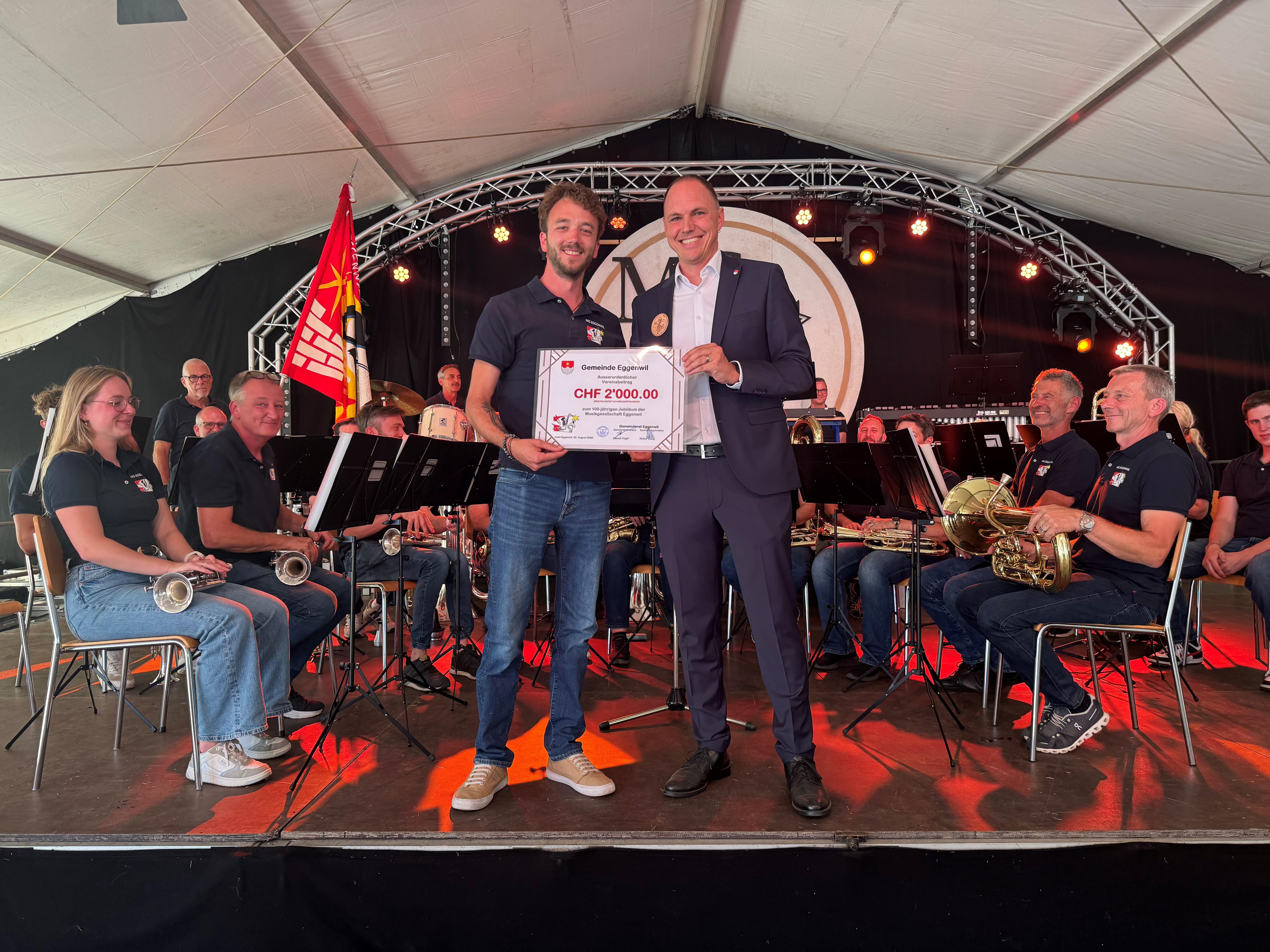 Gemeindeammann Pascal Vogel überreichte im Anschluss an seine wertschätzende Rede der Musikgesellschaft Eggenwil, vertreten durch Yannick Meier, als Dank und Anerkennung einen «Scheck» über 2000 Franken als ausserordentlichen Vereinsbeitrag. Bild: Walter Bürgi/22.08.2026